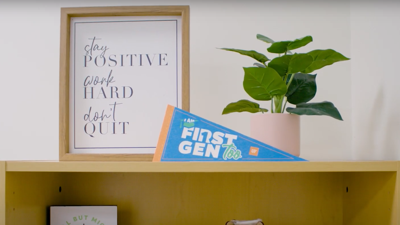 A First-Gen Too pennant displayed on a shelf with a plant and a framed quote "Stay Positive, Work Hard, Don't Quit".